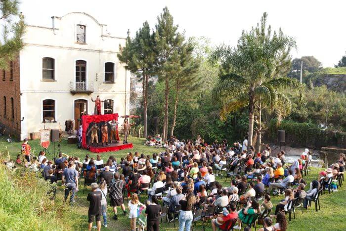 Pessoas sentadas em frente a pequeno palco com apresentação teatral no Moinho da Cascata