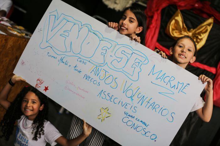 Crianças segurando cartaz de venda de macramê